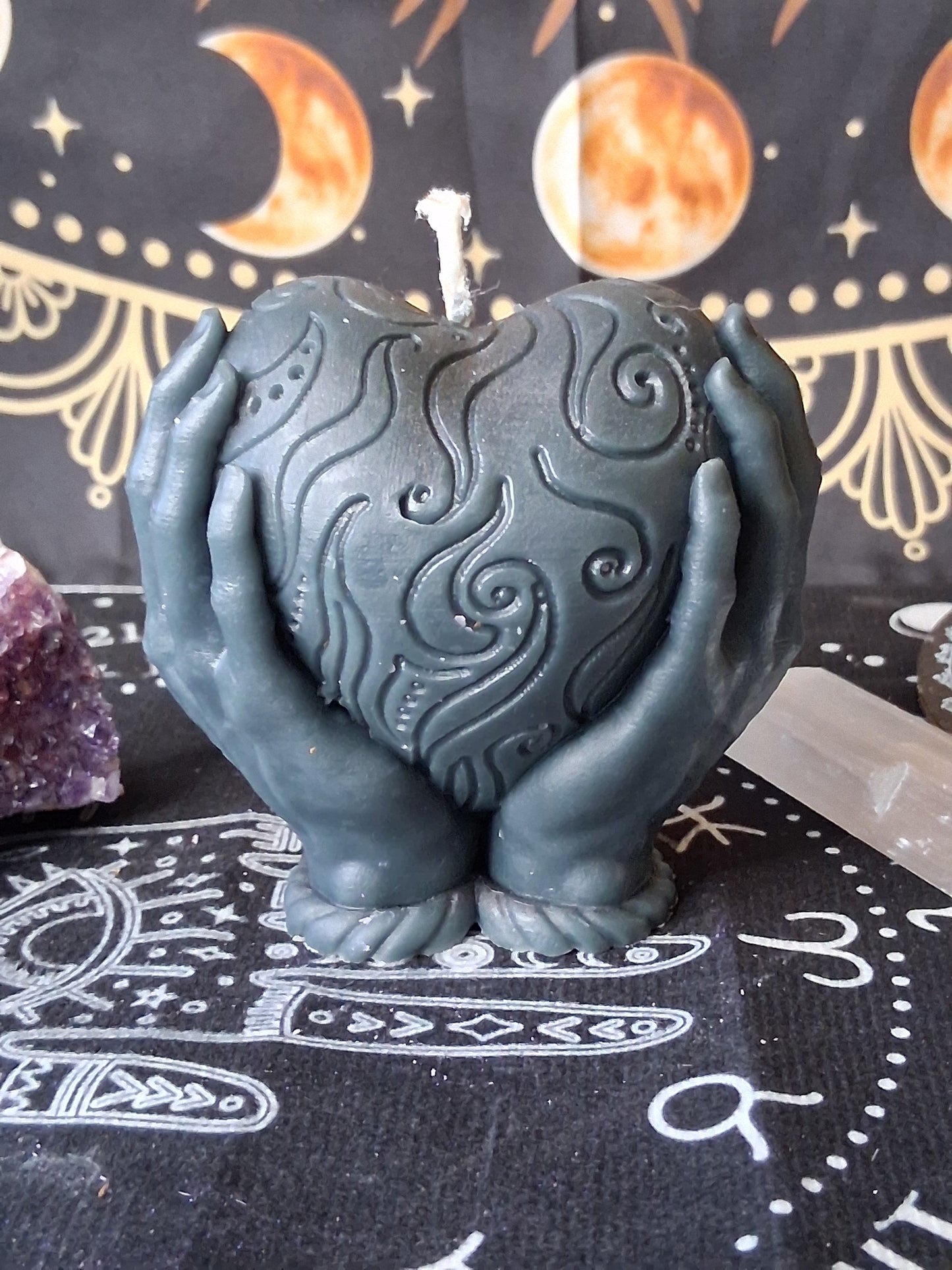 Sacred Moon Candle Co gray heart in hands candle with intricate swirling patterns on black cloth with celestial designs