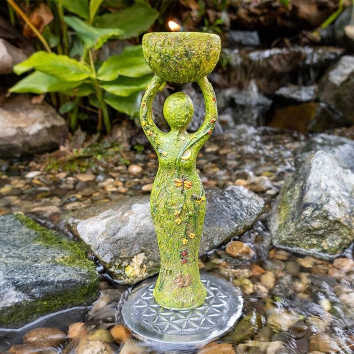 Green goddess altar statue holding a mossy bowl candle on a carved circular base by a rocky stream