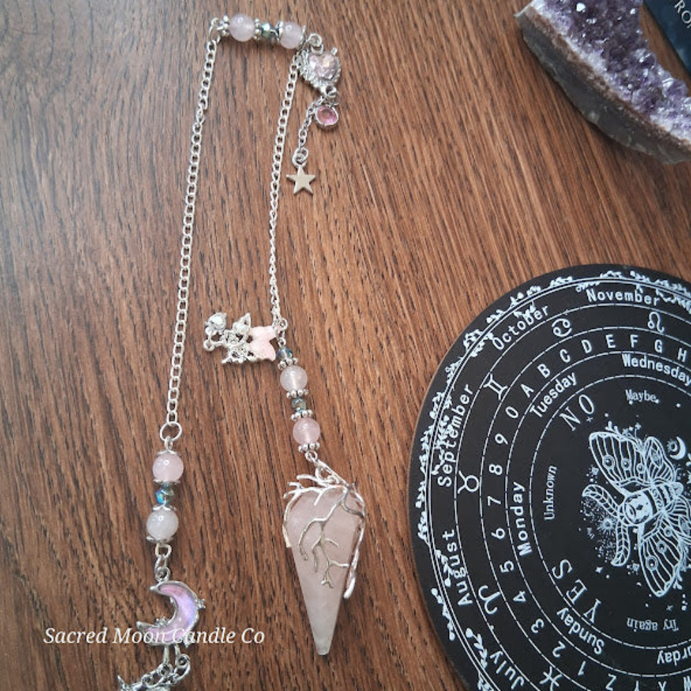 Sacred Moon Candle Co rose quartz pendulum for divination with silver chain and charms on wooden surface