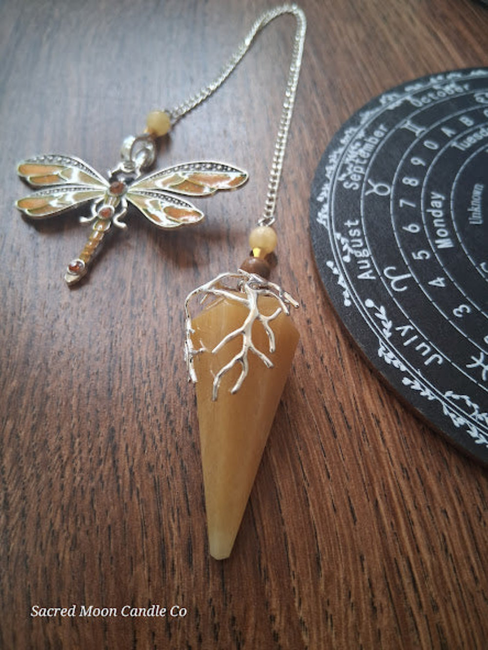 Golden healer quartz pendulum with silver dragonfly charm on wooden surface near black divination board