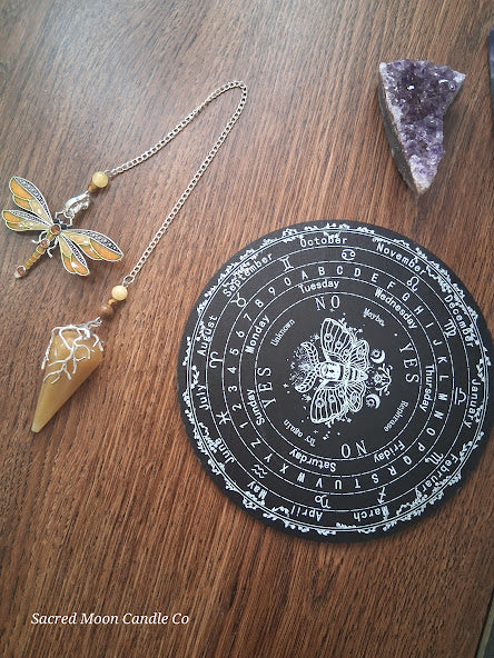 Golden healer quartz pendulum with dragonfly charm on chain beside black divination board and amethyst crystal