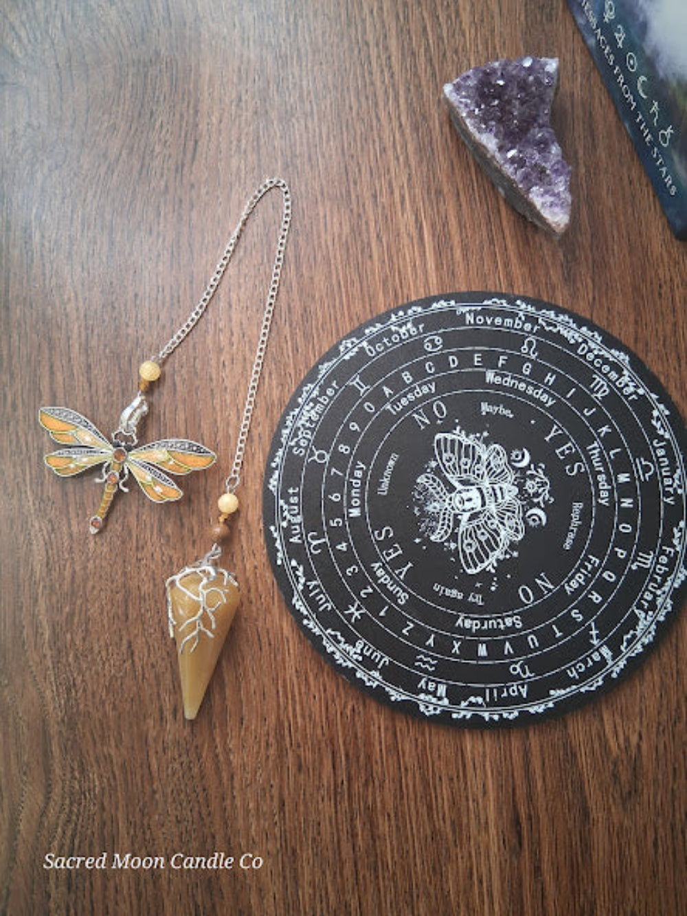Golden healer quartz pendulum with dragonfly charm next to divination board and amethyst crystal by Sacred Moon Candle Co