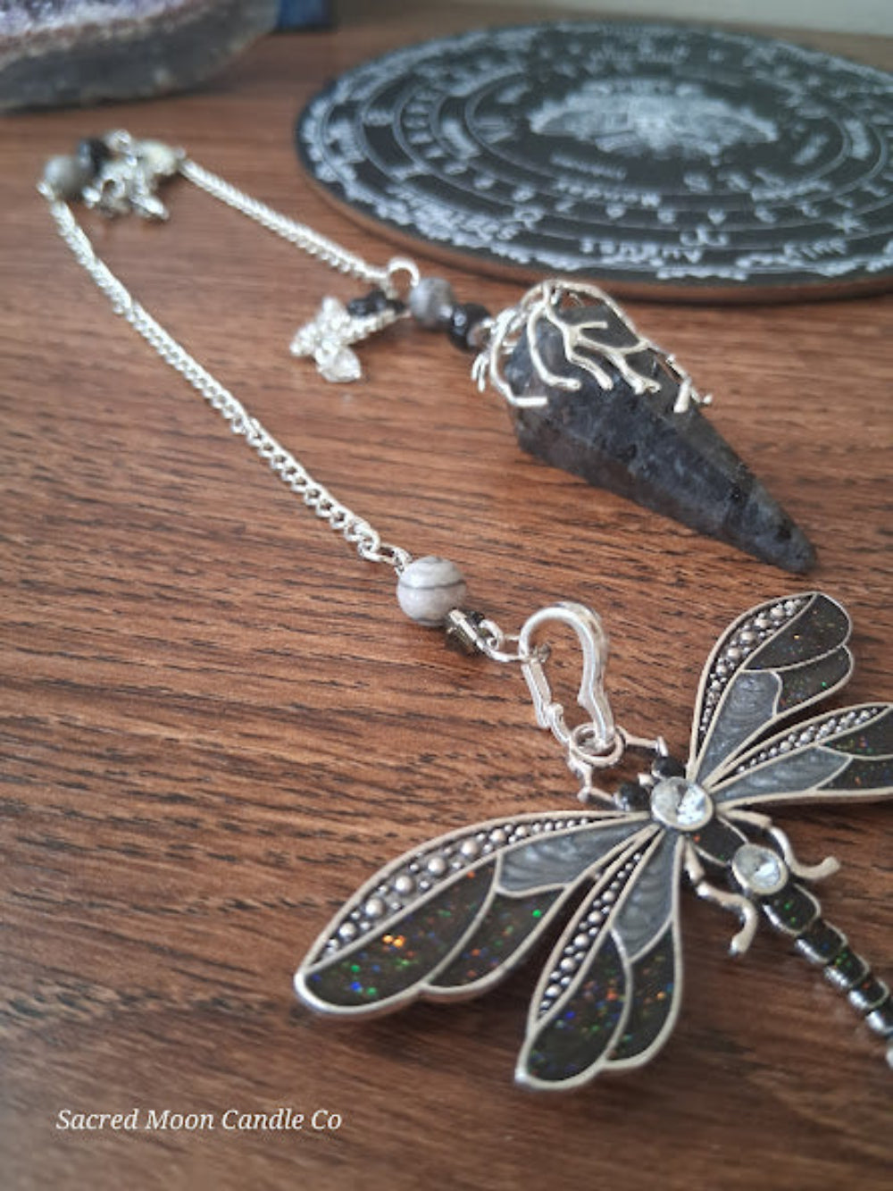 Larvikite moon pendulum with a decorative dragonfly charm on a wooden surface from Sacred Moon Candle Co