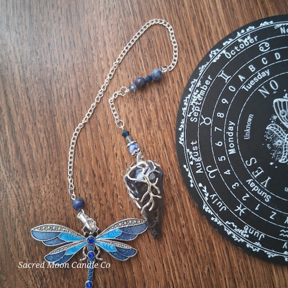 Sodalite and lapis lazuli pendulum with dragonfly design on a wood surface next to a black circular calendar