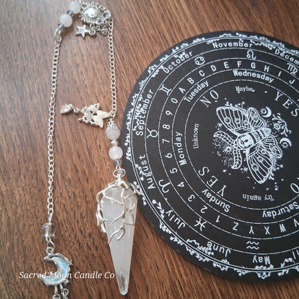 Selenite moon pendulum with silver wire wrap and chain next to black divination board on wooden surface