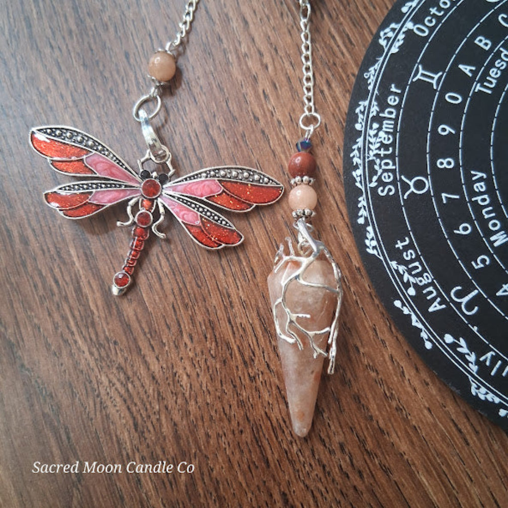 Sunstone dragonfly pendulum for divination with silver chain and decorative dragonfly charm on wooden surface