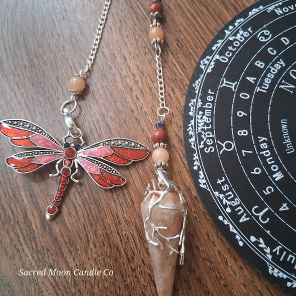 Sunstone dragonfly pendulum for divination with red dragonfly charm and wrapped stone on a wooden surface