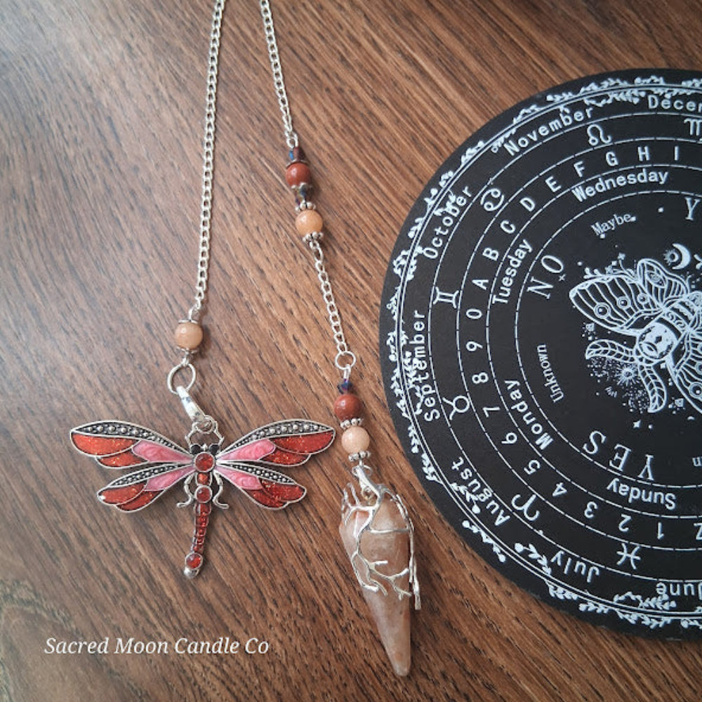 Sunstone dragonfly pendulum for divination with red dragonfly charm and crystal point on wood background