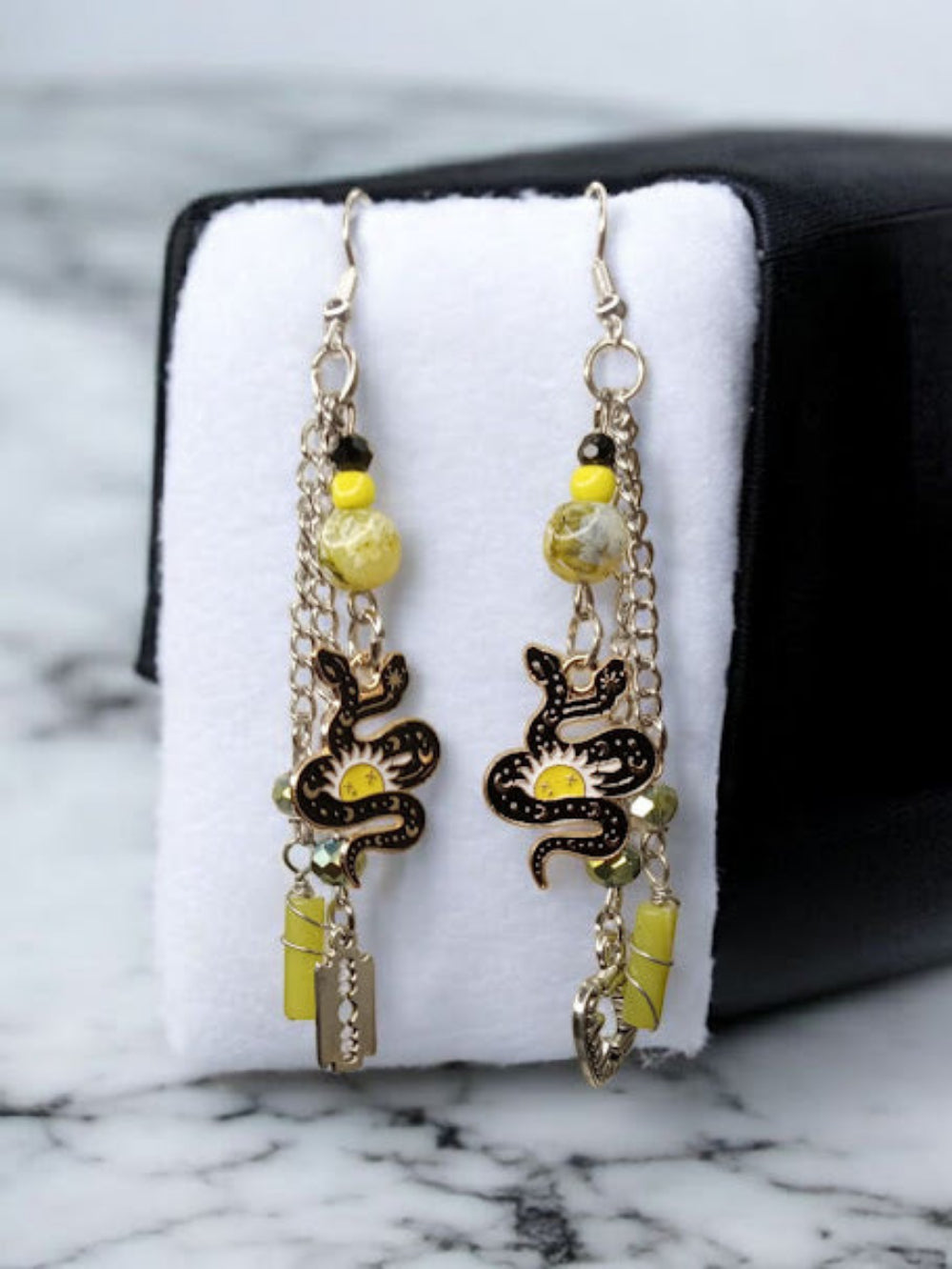 Sacred Moon Candle Co snake charm earrings with yellow beads and silver chains hanging on white display