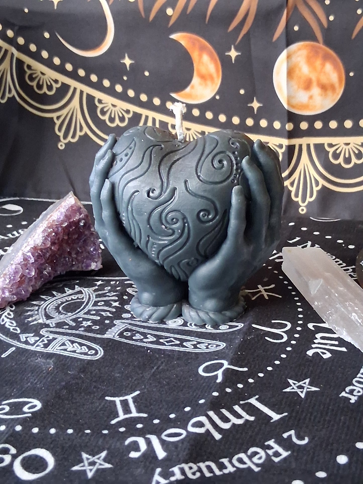 Sacred Moon Candle Co heart in hands candle in dark gray with intricate swirl design surrounded by crystals