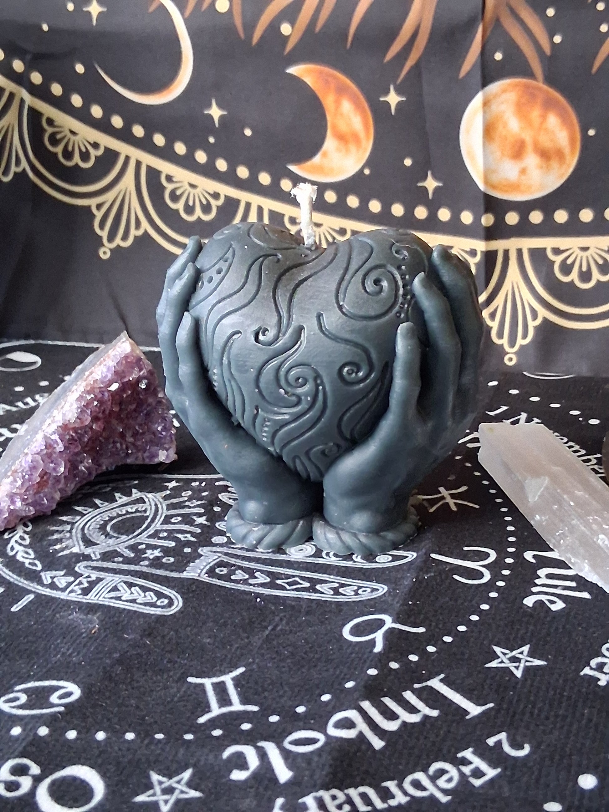 Sacred Moon Candle Co heart in hands candle with intricate swirl design in dark gray on mystical cloth background