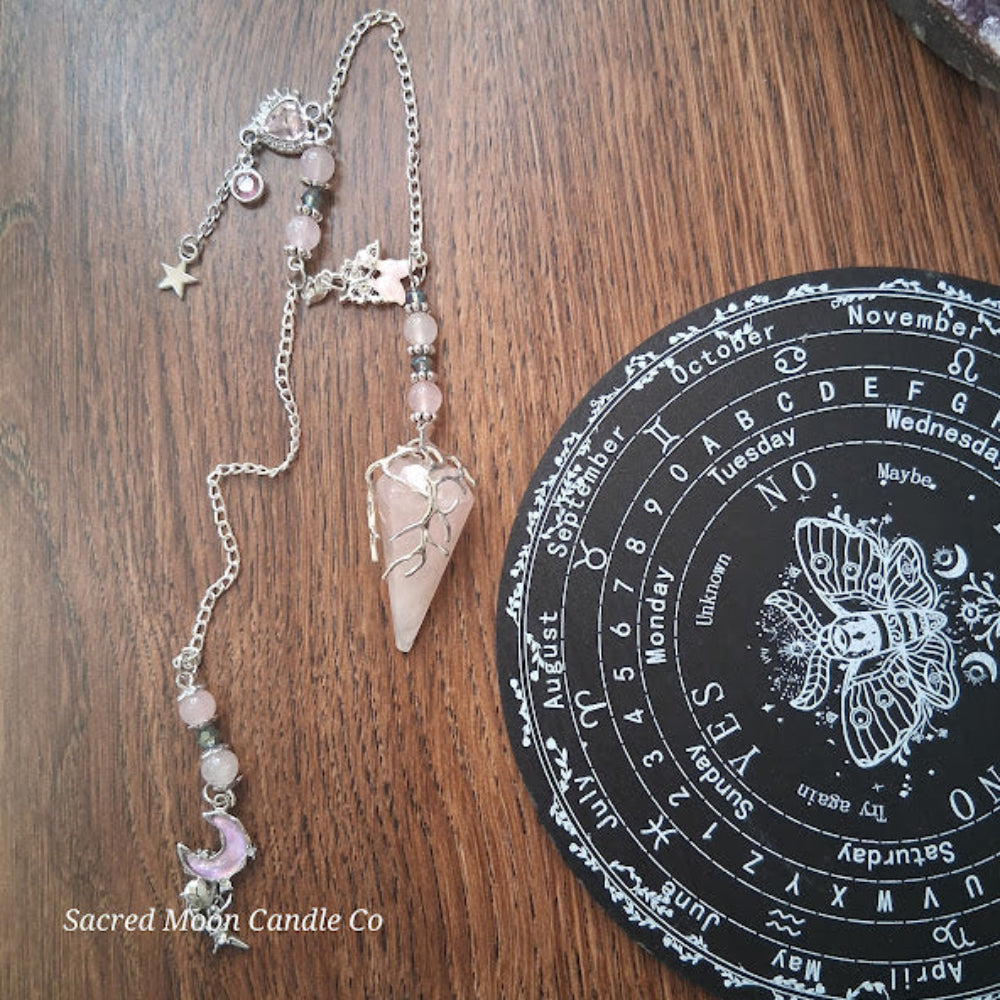 Rose quartz pendulum for divination with silver chain and celestial charms on wooden surface next to black divination board
