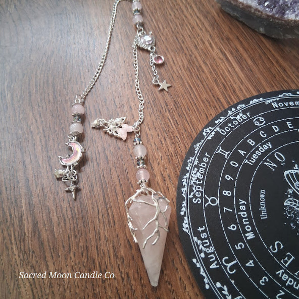 Rose quartz pendulum for divination with silver chain and charms on wooden surface by Sacred Moon Candle Co