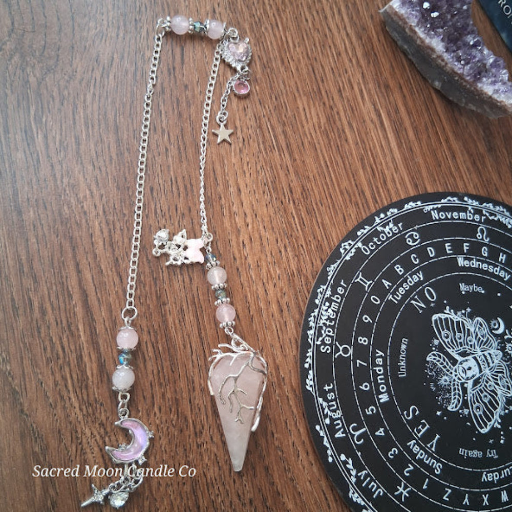 Rose quartz pendulum for divination with silver chain and moon charm on wooden surface beside divination board