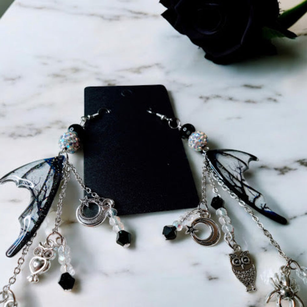 Witches road batwing earrings with intricate silver charms and black and clear beads on a marble surface