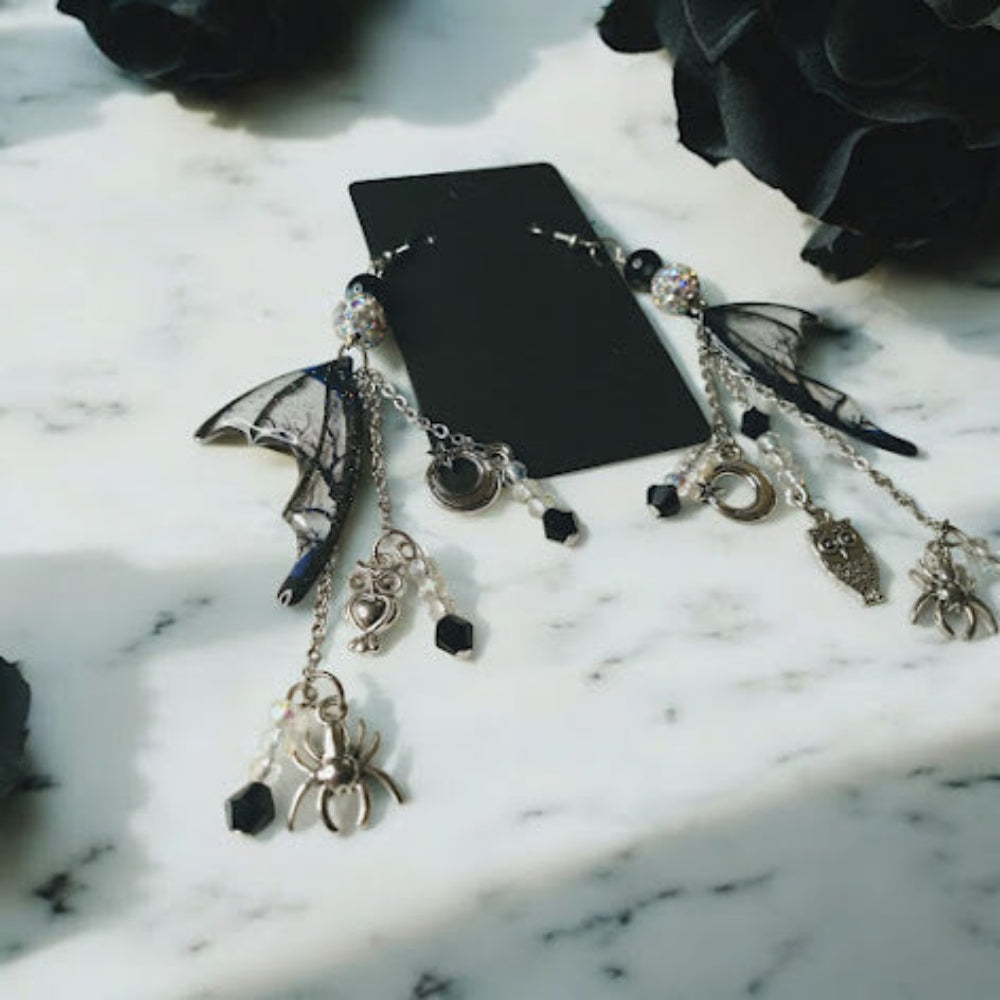 Witches road batwing earrings with black beads and silver charms displayed on marble surface