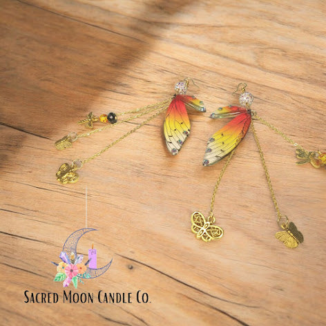 Sacred Moon Candle Co firefly fairy earrings with colorful wing design and gold charm dangles on wooden background