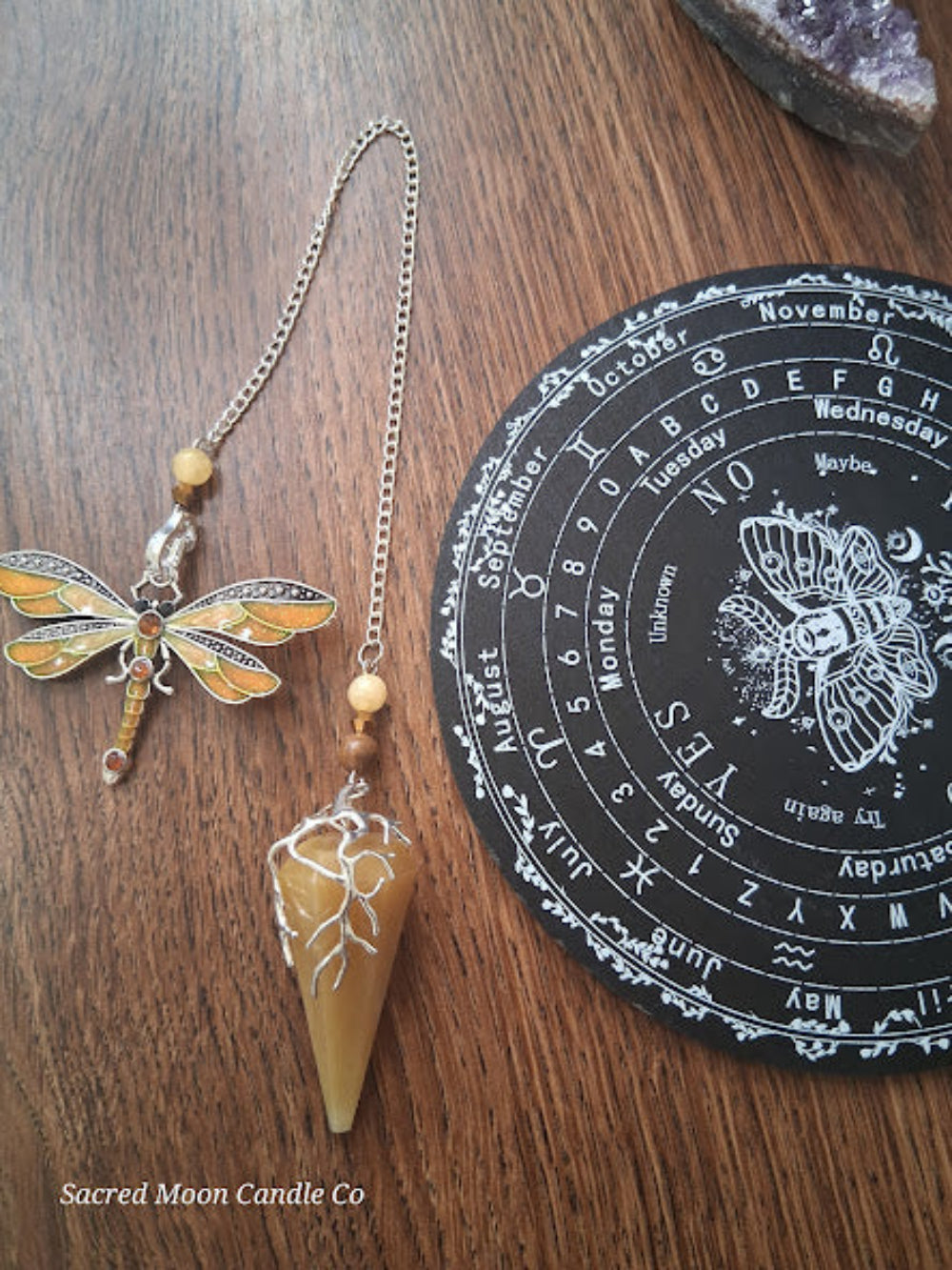 Golden healer quartz pendulum with decorative dragonfly charm and chain on wooden surface near black divination board