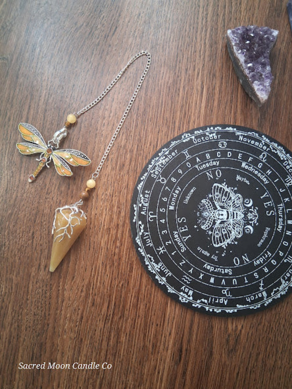 Golden healer quartz pendulum with dragonfly charm next to a black divination board and amethyst crystal