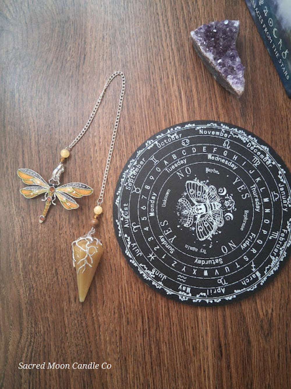 Golden healer quartz pendulum with dragonfly charm next to a black divination board and amethyst crystal on wooden surface