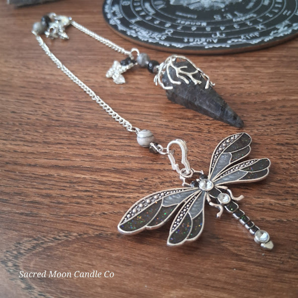 Larvikite moon pendulum with silver dragonfly charm and chain on wooden surface from Sacred Moon Candle Co