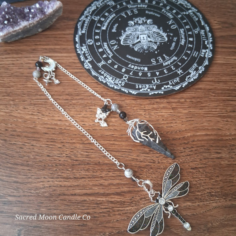 Larvikite moon pendulum with silver dragonfly charm and black stone on wooden surface from Sacred Moon Candle Co