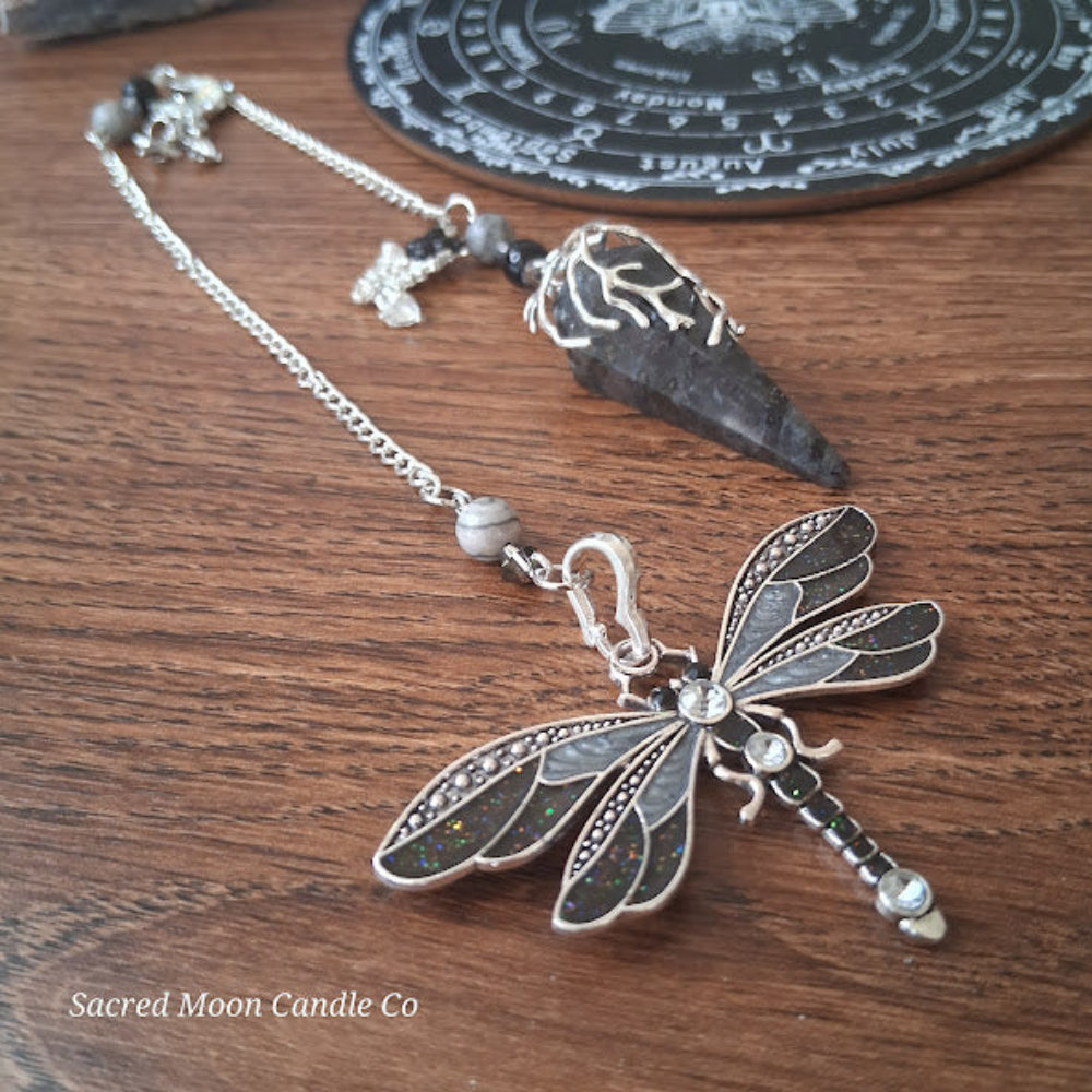 Larvikite moon pendulum with intricate silver dragonfly charm on a wooden surface by Sacred Moon Candle Co