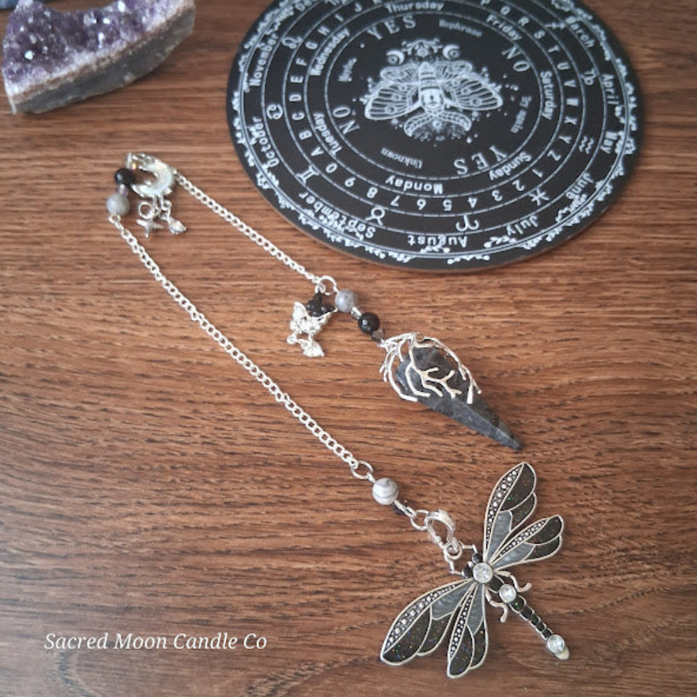 Larvikite moon pendulum with silver dragonfly charm and chain on wooden surface for altar tools