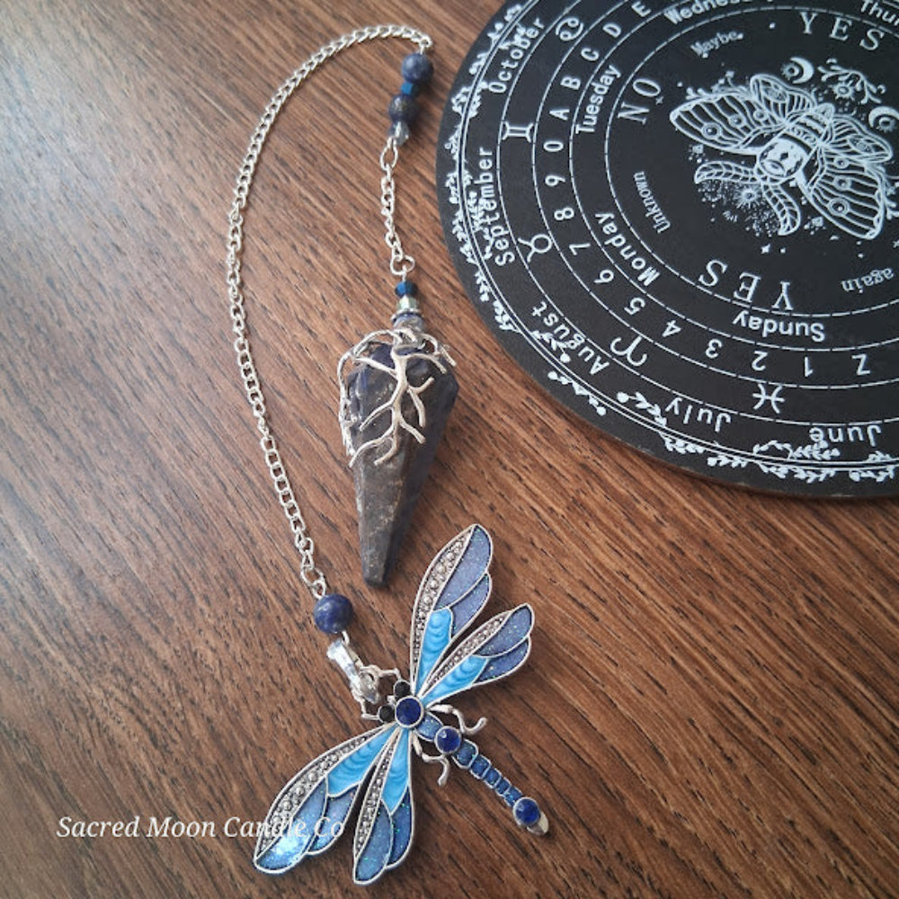 Sodalite and lapis lazuli pendulum with blue dragonfly charm and silver chain on wooden surface near black calendar board