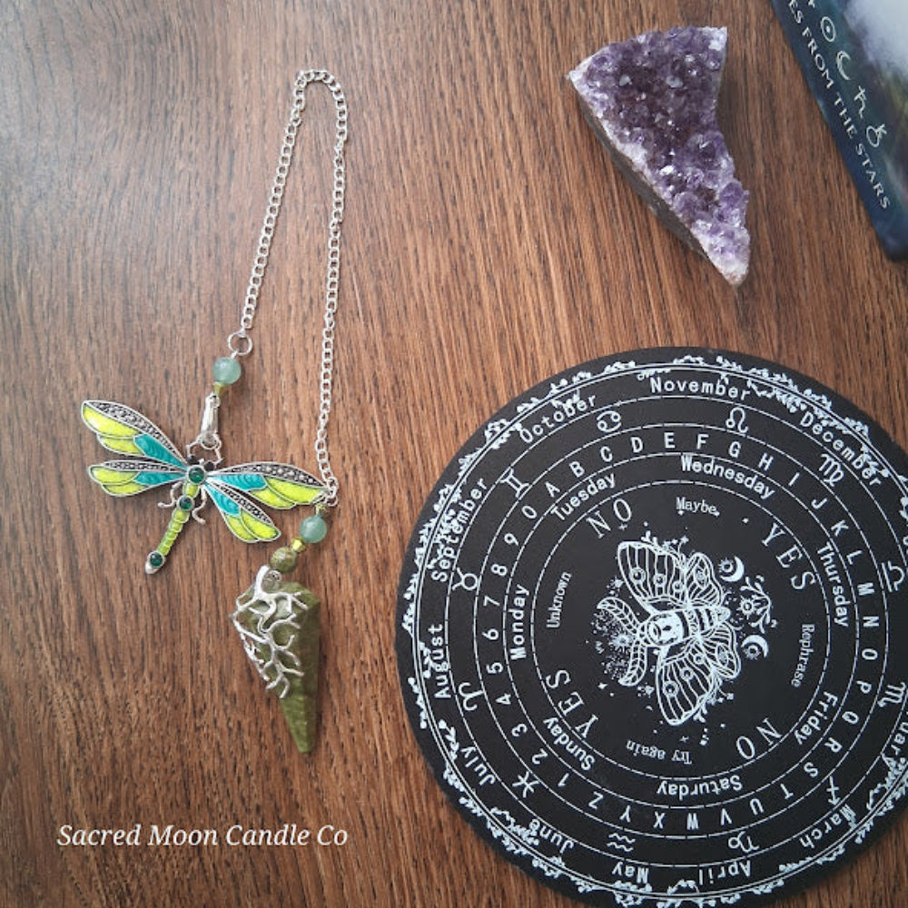 Unakite amazonite dragonfly pendulum with green stone and yellow blue wings on wood surface with divination board and amethyst crystal