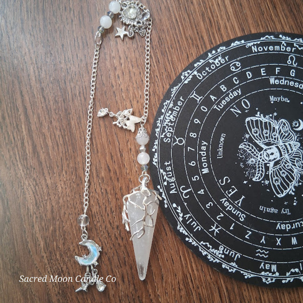 Selenite moon pendulum with silver chain and charms from Sacred Moon Candle Co on wooden surface
