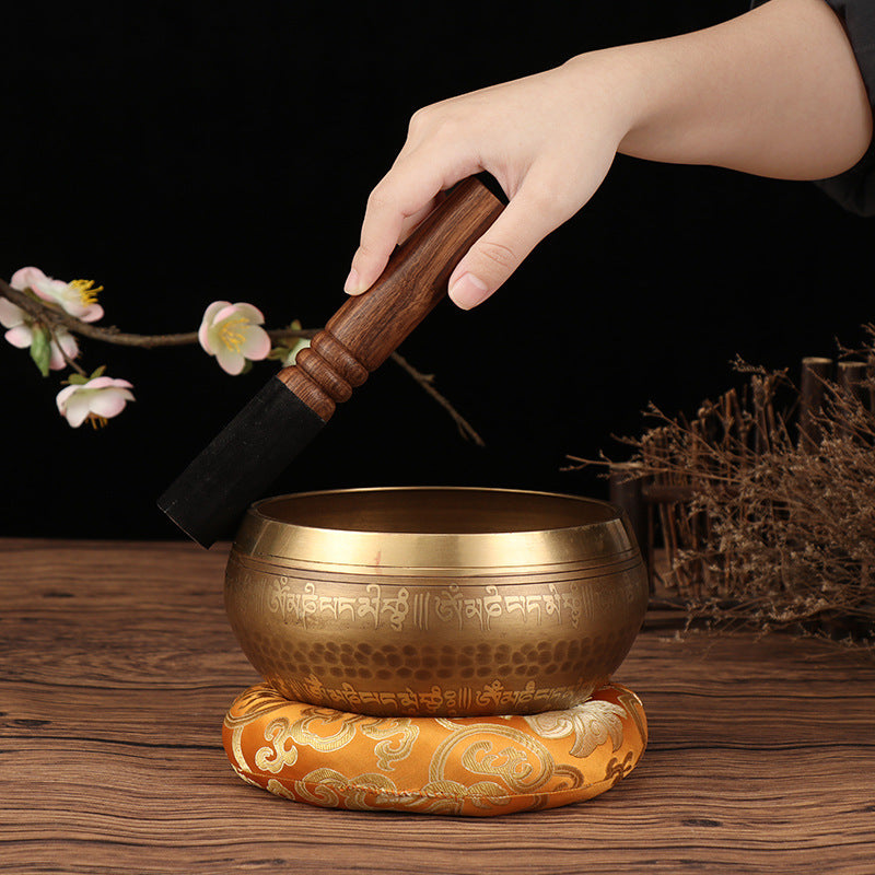 Tibetan Buddha mantra bowl with wooden striker and ornate cushion on wooden surface by Sacred Moon Candle Co